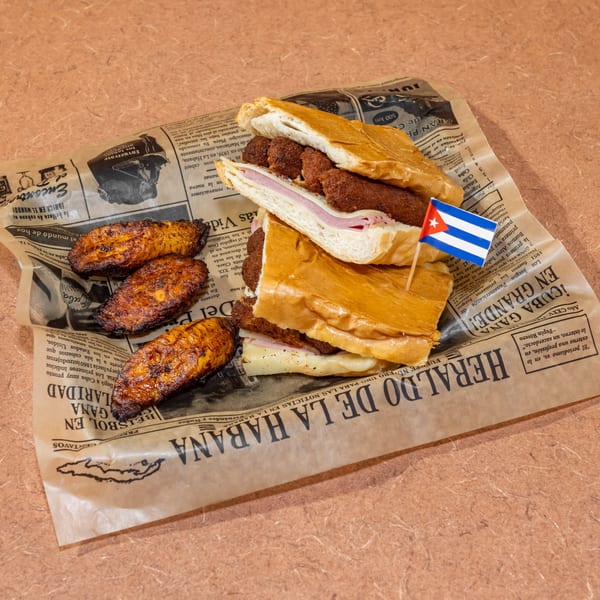 Cuban sandwich with croquettes and ham, served with fried plantains on newspaper-style paper.