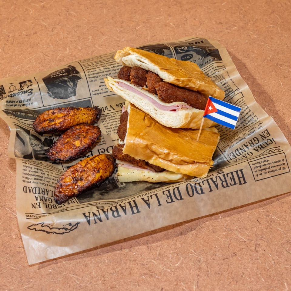 Cuban sandwich with croquettes and ham, served with fried plantains on newspaper-style paper.