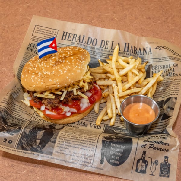 A Cuban Diner burger with a Cuban flag pick, fries, and dipping sauce on newspaper-print paper.