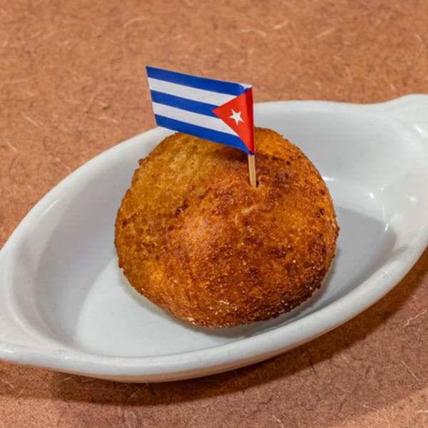 A golden-brown Cuban croquette with a small Cuban flag toothpick, served in a white dish.