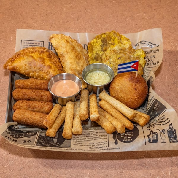 A Cuban appetizer platter with empanadas, croquettes, tostones, and dipping sauces on newspaper-print paper.